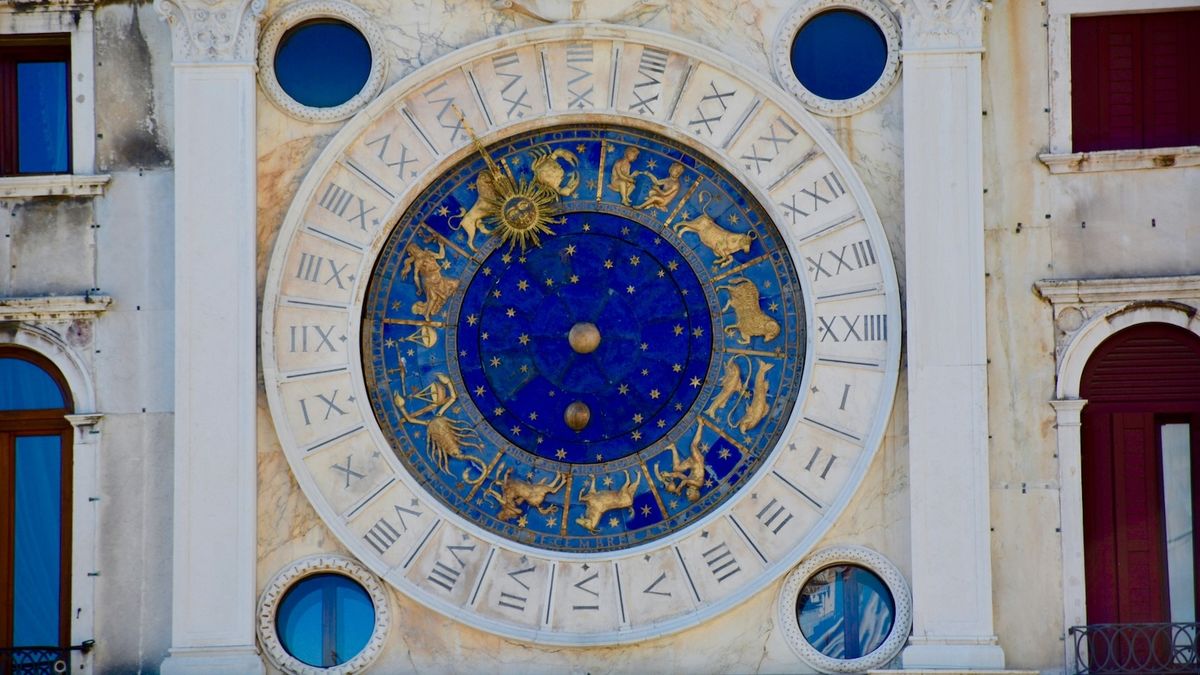 The blue and gold astronomical clock of St Mark's Clocktower in Venice: an outer ring of Roman numerals cut into pale marble, and an inner dial of deep lapis blue scattered with gold stars, ringed by the twelve zodiac figures in gilt relief &mdash; the crab of Cancer, the lion of Leo, the bull of Taurus, the scorpion of Scorpio among them. A large golden sun with a human face and radiating rays points to Leo.